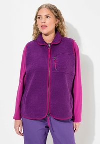 Lila Fleece-Weste mit Reißverschluss, hohem Kragen und einer Brusttasche. Kombiniert mit einem langärmeligen pinken Oberteil und lila Hosen.