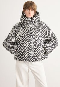 Giacca puffer a fantasia bianco e nero con collo alto e cappuccio, caratterizzata da disegni geometrici a zigzag e polsini elasticizzati.