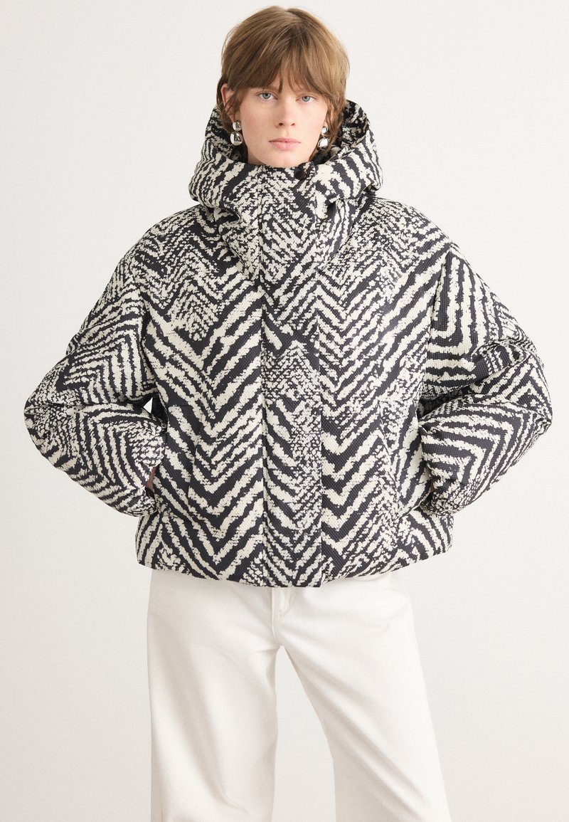 Giacca puffer a fantasia bianco e nero con collo alto e cappuccio, caratterizzata da disegni geometrici a zigzag e polsini elasticizzati.