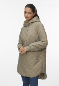 Veste à capuche beige matelassée avec un col haut, des manches longues et des poches latérales. Présente une surface texturée et un ourlet arrondi.
