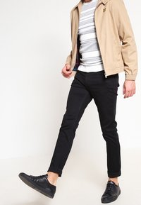 Chaqueta ligera beige, suéter a rayas blanco y gris, pantalones negros ajustados y zapatillas negras. Modelo caminando con un pie adelante.