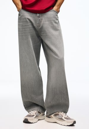 Personne portant un jean gris ample avec les mains dans les poches et des baskets beiges et grises sur fond blanc.