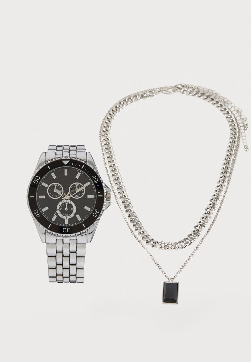 Silbernes Metall-Armband mit schwarzem Zifferblatt und Chronographenfunktionen, kombiniert mit einer silbernen Kettenhalskette, die einen rechteckigen schwarzen Anhänger trägt.