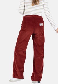 Roestkleurige corduroybroek met wijde pijpen, met achterzakken en een decoratieve patch aan de rechterkant. De stof heeft een geribbelde textuur.