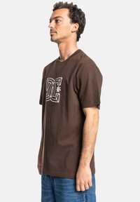 Camiseta de algodón marrón con cuello redondo, que presenta un estampado gráfico blanco en el pecho. Corte relajado con mangas cortas y dobladillo estándar.