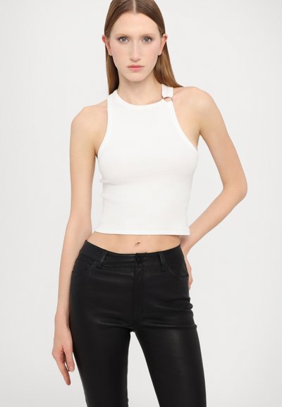 B�ílý žebrovaný crop top s vysokým výstřihem a postranním detailním okem, v kombinaci s černými vypasovanými kalhotami. Jednoduché pozadí zdůrazňuje outfit.