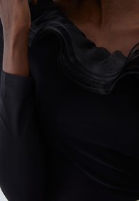 Top nero con un colletto arricciato morbido e trasparente e maniche lunghe. Il tessuto appare liscio e leggermente elastico. Gli accenti mettono in risalto il design del collo.