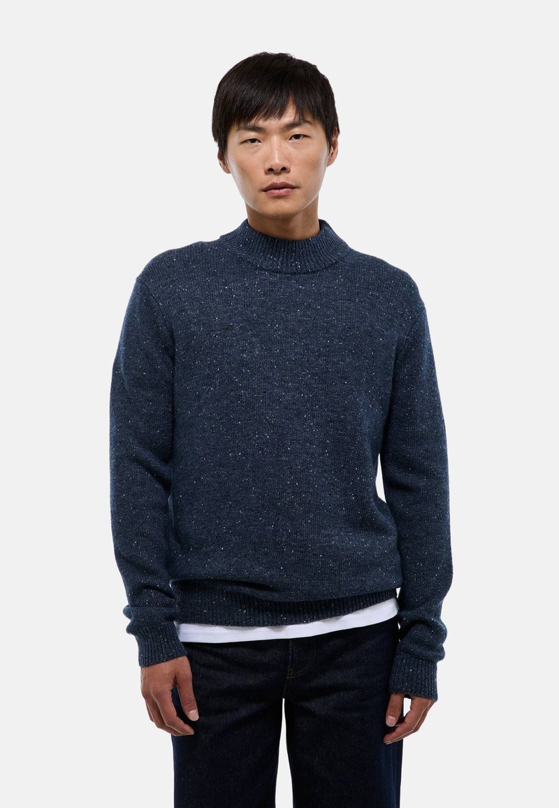 Pull en maille bleu marine avec un col montant, présentant une finition texturée, des manches longues, et des bords côtelés au niveau des poignets et de l'ourlet. Porté par-dessus une chemise blanche.