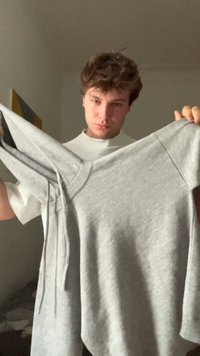 Jonge man met krullend haar die een grijze sweatshirt vasthoudt, uitgestrekt, binnen met een muur in neutrale kleuren en een abstract schilderij op de achtergrond.