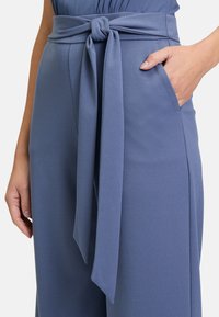 Pantalon bleu clair avec une large taille et un nœud noué à l'avant ; dispose de poches latérales et d'une texture de tissu lisse.