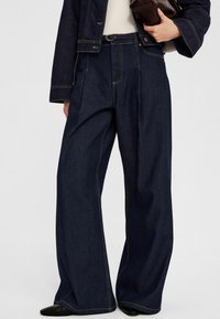 Mörkblå vida denimbyxor med framveck, kontrasterande sömmar, hög midja och släta framfickor. Matt svarta spetsiga skor.