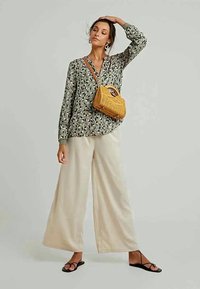 Mulher vestindo uma blusa de manga longa com padrão, calças largas bejas, sandálias pretas, segurando uma bolsa de vime, em pé contra um fundo claro e simples.
