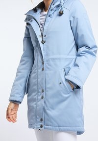 Manteau bleu clair longueur genoux avec fermeture éclair à l'avant, boutonnière et capuche. Dispose de deux poches latérales et de poignets côtelés. Tissu doux et texturé.
