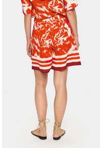 Rdeče kratke hlače z valovitim vzorcem, ki imajo širok pas in rob z bordo in smetanastimi črtami. Nosijo se z bež čipkastimi natikači.