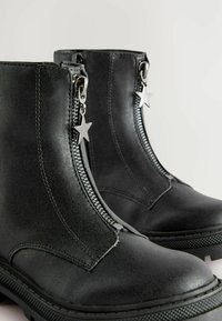 Bottines noires avec des fermetures éclair à l'avant, dotées de tirettes en forme d'étoile en argent, en cuir texturé, et avec une semelle épaisse sur fond blanc.