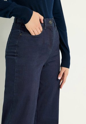 Henkilö, joka pukeutunut tummansinisiin, korkeavyötäröisiin leveälahkeisiin farkkuihin ja tummansiniseen neulepuseroon, vasen käsi edessä taskussa ja oikea käsi rentona sivulla.