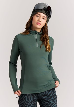 Frau trägt ein grünes Zip-up-Oberteil, schwarze Mütze mit Skibrille und schwarze und blaue Zebramusterhose, posiert mit Händen am Saum.