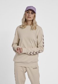 Felpa beige con maniche lunghe, decorata con motivi a zigzag marroni sulle maniche, abbinata a pantaloni coordinati e un cappellino lavanda.