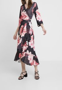Blommig omslagsklänning i svart med rosa och röda blommor, med lång ärm, knytmidja och volangkant. Svarta sandaler med remmar.