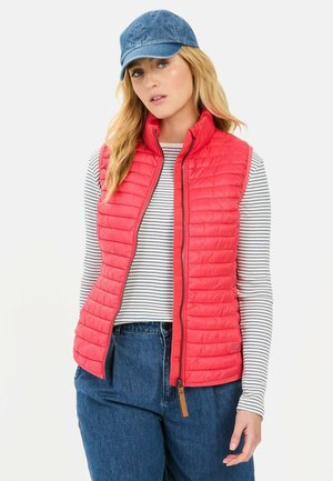 Red puffer vest with quilted texture, zip front, and high collar, paired with a striped long-sleeve shirt and denim pants. Accented with a blue cap.
