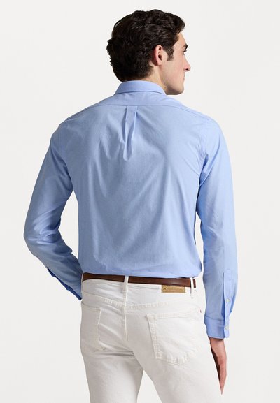 Gaiši zils krekls ar aizmugures plisējumu, garām piedurknēm un gludu tekstūru, kombinēts ar baltām biksēm un brūnu ādas jostu.