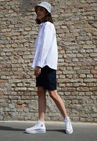 White button-up shirt with long sleeves, black shorts, and white high-top sneakers; floral-patterned bucket hat. Set against a brick wall.