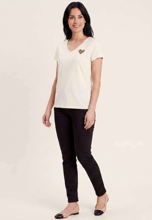 Femme debout portant un haut blanc à manches courtes en V avec un cœur imprimé léopard, un pantalon noir et des ballerines beiges avec des bouts noirs.