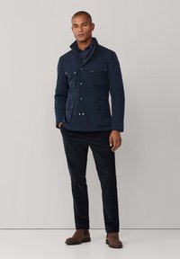 Mand iført en marineblå jakke med fire forlommer, mørke bukser og brune støvler, stående mod en ensfarvet lys baggrund.