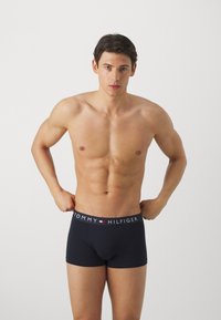 Boxers navy avec une ceinture logo TOMMY HILFIGER. Tissu doux et extensible ; design ajusté ; longueur jusqu'au milieu de la cuisse. Le modèle pose les mains sur la ceinture.