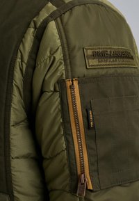 Veste matelassée verte avec une finition texturée, dotée d'une poche zippée, d'un écusson logo brodé et de quincaillerie dorée contrastante.