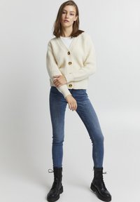 Creme strikket cardigan med V-udskæring, der har tre store brune knapper. Kombineret med blå skinny jeans og sorte snørebåndsstøvler.