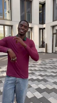 Uomo sorridente che indossa una camicia a maniche lunghe bordeaux e jeans chiari, gesticola con le mani mentre si trova su un pavimento decorato all'esterno di un edificio moderno.