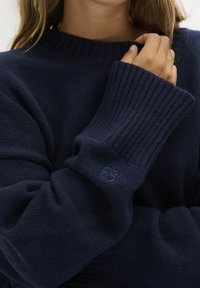 Navyblauw gebreid sweater met geribde manchet en kraag. Bevat een klein logo op de mouw. Zachte textuur en relaxte pasvorm.