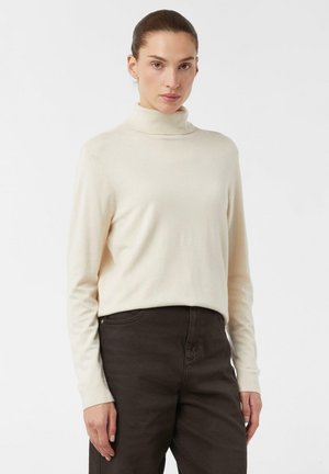 Woman with brown hair in a bun wearing a cream turtleneck sweater and dark brown pants against a plain white background.