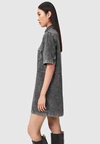 Pitkähiuksinen nainen, jolla on lyhythihainen, polvenpituinen haalistunut musta farkkumekko ja mustat puolipitkät saappaat, seisoo sivuttaisnäkymässä.