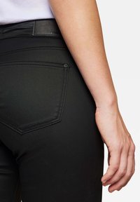 Schwarze, eng anliegende Jeans mit glatter Textur, ausgestattet mit einer Gesäßtasche mit Knopf und einem flachen, markierten Bund. Nahaufnahme der Hand, die auf der Hüfte ruht.