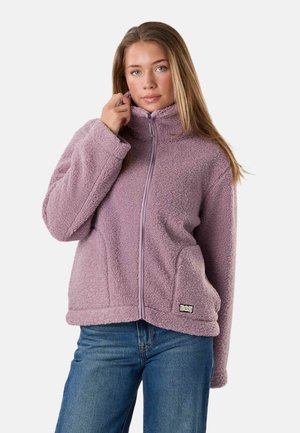 Kvinde iført en lavendelfarvet fleecejakke med lynlås og blå jeans, der holder jakke kraven med den ene hånd, står mod en ensfarvet baggrund.