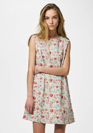 Tânără cu păr lung și blond, purtând o rochie lejeră fără mâneci cu model floral roșu, albastru și galben, stând pe un fundal alb.