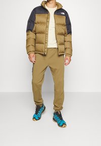 Giubbotto imbottito con corpo beige e spalle nere, maglietta a girocollo beige, pantaloni jogger oliva e sneaker blu-neri con dettagli gialli.