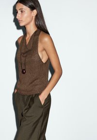 Top senza maniche in maglia marrone con scollo a cowl, abbinato a pantaloni larghi verde oliva con tasche laterali, presentato su uno sfondo semplice.