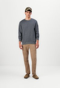Felpa grigia a maglia con polsini a costine, pantaloni cargo beige con più tasche e scarpe slip-on marroni. Berretto oliva con dettaglio testuale.