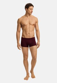 Tmavě červené boxerské trenýrky z měkké látky, s páskem v pase a přiléhavým střihem. Model stojí bosý, mírně otočený do strany.