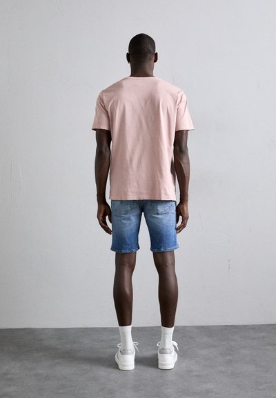 Camiseta de algodón rosa, ajuste relajado, combinada con pantalones cortos de mezclilla azul. El modelo lleva zapatillas deportivas blancas y calcetines texturizados, visto desde atrás.