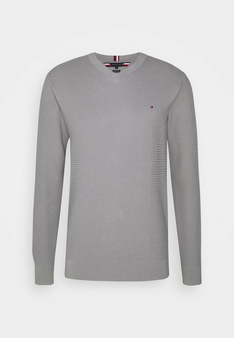 Pull à manches longues gris clair avec col en V, motif en tricot texturé et petit logo rouge, blanc et bleu sur le côté gauche de la poitrine.