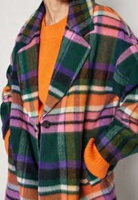 Kvinde iført en farverig skotskternet frakke med grøn, pink, lilla og orange over en lys orange strikketrøje, hænderne gemt i frakkens lommer.