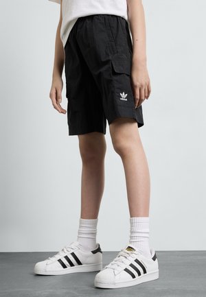 Schwarze Cargo-Shorts aus einem strukturierten Stoff, mit einem adidas-Logo auf der Vorderseite. Kombiniert mit weißen Sneakers mit schwarzen Akzenten und gerippten Socken.