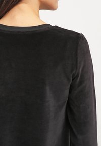 Top de terciopelo negro con una textura suave y mangas largas. El escote está terminado con un simple ribete oscuro, mostrando una caída suave en la parte trasera.