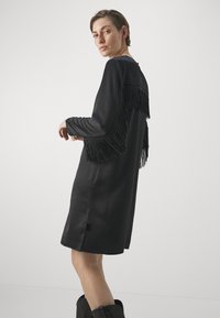 Abito nero con maniche lunghe, caratterizzato da dettagli frangiati sulla schiena e sulle spalle. Tessuto morbido, taglio al ginocchio, design semplice.