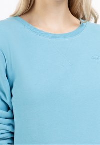 Ljusblå sweatshirt med rund halsringning, ribbade ärmslut och subtilt broderad paraplydetalj på bröstet. Mjuk, texturerad tyg.