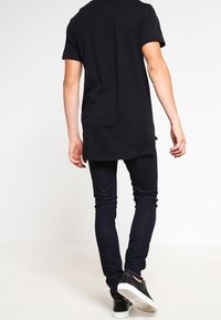 Svart t-shirt med kort ärm och längre bakstycke, tillsammans med mörka slim-fit jeans och svarta sneakers med vita sulor; avslappnad och väl avpassad stil.